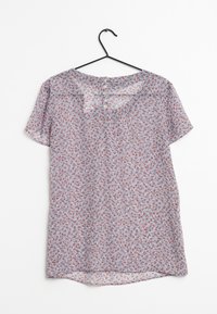 Blusa floreale a maniche corte con piccoli fiori rossi e blu, colletto rotondo e quattro bottoni sul retro, appesa a un gancio nero.