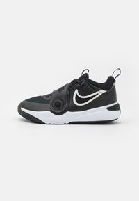 Chaussure de sport noire avec une tige en textile, des accents blancs, un détail de logo circulaire, des superpositions texturées et une semelle en caoutchouc blanche.