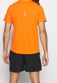 Camiseta atlética corta de manga en color naranja con una textura acanalada y detalle reflectante en la parte posterior, combinada con pantalones cortos negros.