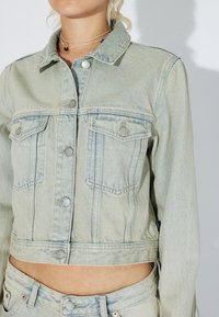 Mujer con chaqueta corta de denim azul claro con cierres de botones y vaqueros de talle alto a juego, mostrando el abdomen y con collares en capas.