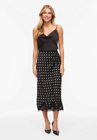 Camisole en satin noir et jupe à pois avec ourlet en dentelle. La jupe présente des pois blancs et une silhouette épurée, complétée par des talons noirs.