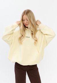 Overgrote gele hoodie met een voorzak, voorzien van groene geborduurde tekst "MIRONS" op de borst, gecombineerd met bruine corduroy broek.