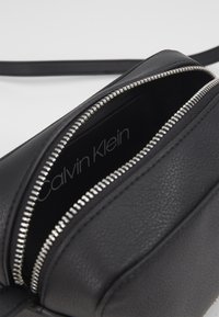Černá kožená kabelka crossbody se zipovým uzávěrem, s hladkou texturou a stříbrným zipem. Uvnitř je viditelné logo "Calvin Klein".