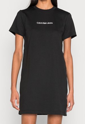 Robe décontractée noire à manches courtes avec le logo "Calvin Klein Jeans" centré sur la poitrine, portée par une personne sur un fond clair uni.