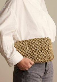 Geweven gouden clutch met een gestructureerd, geknoopt ontwerp, vastgehouden in één hand tegen een wit shirt en grijze jeans. Rechthoekige vorm, compacte maat.