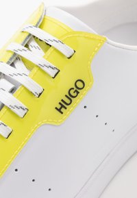 Sneaker bianco con dettagli in pelle verniciata gialla, logo nero "HUGO" e lacci bianchi testurizzati. Presenta perforazioni per la traspirabilità.