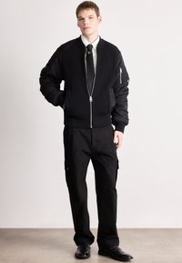 Casaco bomber preto com textura canelada, fecho frontal com zíper e dois bolsos laterais. Combinado com calças pretas largas e sapatos elegantes.