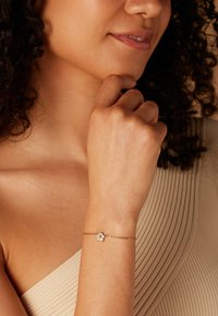 Vrouw met krullend haar die een beige geribde top draagt, toont een delicate gouden armband met een bloemblaadje-vormige bedel om haar pols.