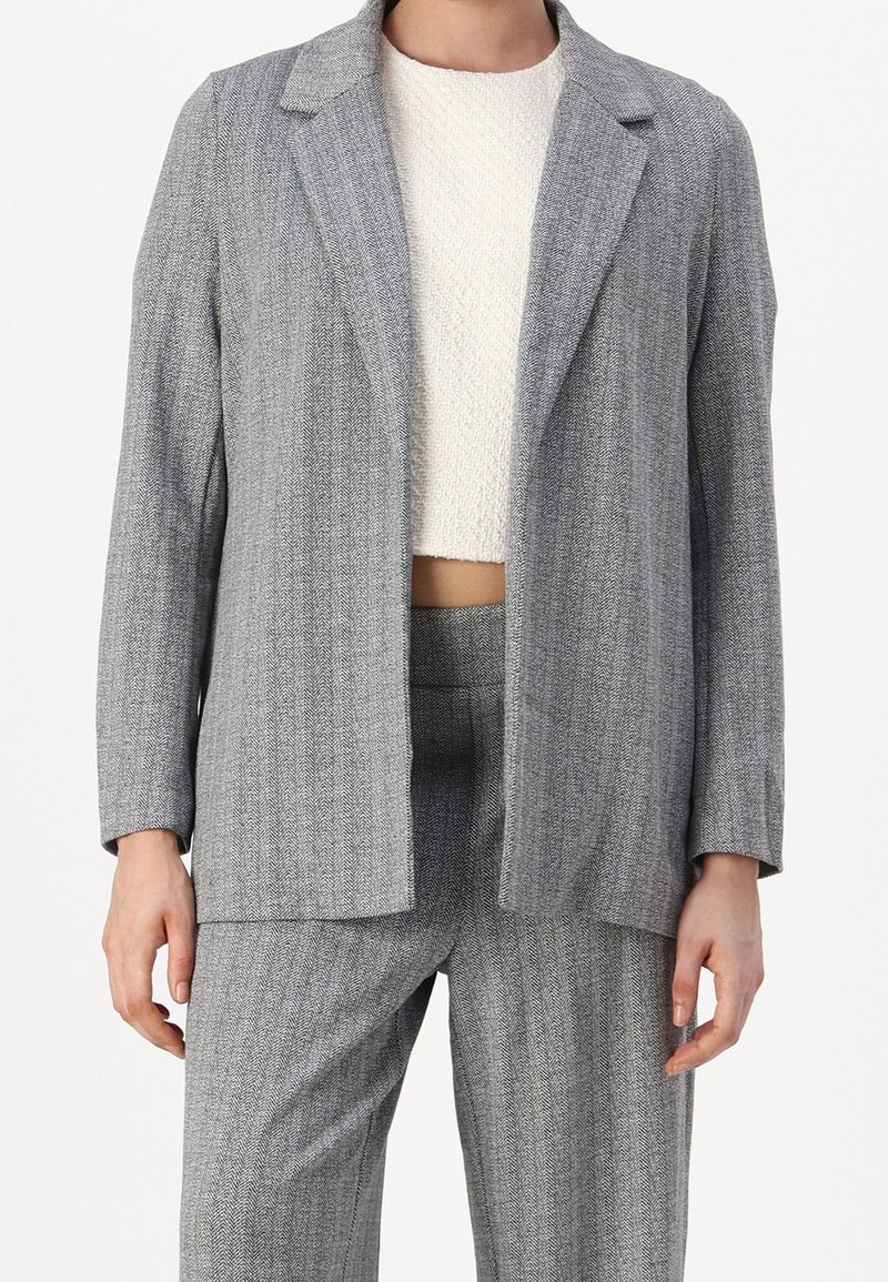 Personne portant un costume gris en chevrons avec un blazer ouvert et un pantalon assorti, associé à un top court blanc texturé.