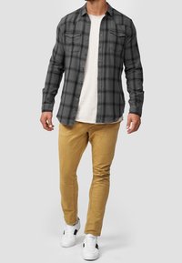 Chemise en flanelle à carreaux gris avec fermeture boutonnée, deux poches poitrine, portée sur un t-shirt blanc à col rond. Associée à un pantalon beige ajusté et des baskets blanches.