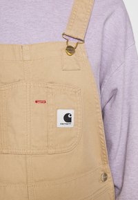 Béžové džínové lacláče s nastavovacími popruhy, zlatým kováním, náprsní kapsou s logem Carhartt, spárované s dlouhým svetrem světle fialové barvy.