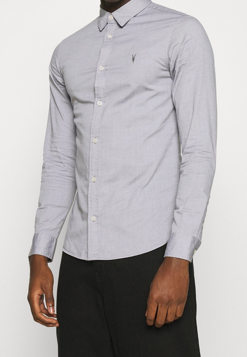 Camicia button-up grigia chiara con colletto classico, maniche lunghe e un piccolo logo ricamato sul petto. Tessuto morbido.