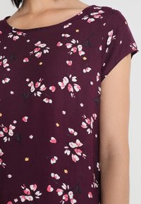 Kobieta ubrana w bordową bluzkę z krótkim rękawem w różowe i białe kwiatowe wzory oraz okrągłym dekoltem.