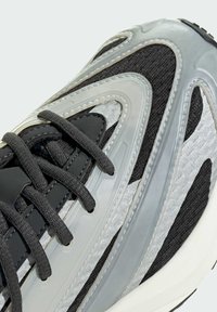 Čierne a sivé tenisky s textúrovaným sieťovým zvrškom, lesklými striebornými akcentmi a zakriveným plastovým prekrytím. Šnúrky sú čierne a pevne zaviazané.