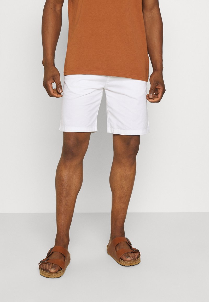 Shorts en coton blanc avec ourlet replié, associés à des sandales en cuir marron avec boucle. Modèle portant un t-shirt marron.