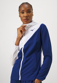 Zila un balta sporta jaka ar rāvējslēdzēju, augstu apkakli, diagonālu sadalījuma dizainu un nelielu logo uz krūtīm. Gluda tekstūra.