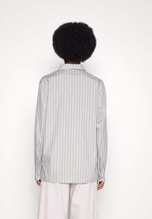 Person med kort krøllet hår iført en grå og hvid vertikalt striped langærmet skjorte og hvide bukser, set bagfra mod en hvid baggrund.