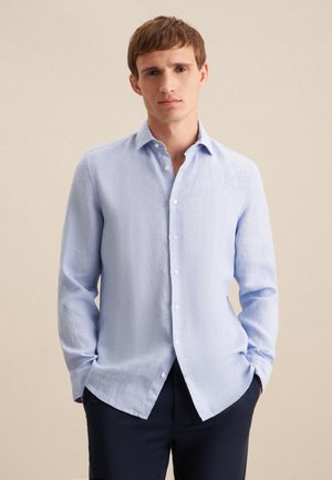 Camicia button-up azzurro chiaro in un tessuto texturizzato, con colletto classico, maniche lunghe e orlo curvo. Indossata con pantaloni scuri.