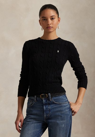 Donna con orecchini che indossa un maglione nero a trecce e jeans blu con una cintura nera intrecciata, in piedi davanti a uno sfondo semplice.