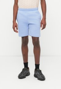 Uomo con pantaloncini in pile azzurro chiaro, calzini neri e scarpe da ginnastica nere in piedi contro uno sfondo bianco.