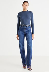 Haut long à manches ribbed gris foncé avec un décolleté en dentelle, associé à un jean droit taille haute bleu et des sandales à talons violettes.