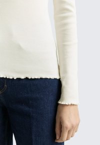 Close-up van een persoon die een geribd, gebroken wit longsleeve draagt met slabruimtes aan de manchetten en zoom, gecombineerd met donkerblauwe denim jeans.