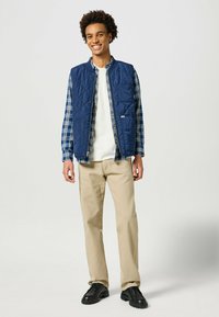 Chaleco acolchado azul marino sobre una camisa de cuadros, combinado con pantalones beige y zapatos negros. El material parece suave con una textura ligera.