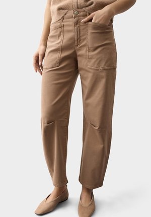 Vrouw draagt beige hoge taillebroek met grote voorzakken en beige platte schoenen, staand tegen een eenvoudige lichte achtergrond.
