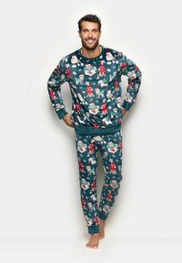 Uśmiechnięty mężczyzna stoi boso w turkusowych piżamach z wzorem świątecznych psów i prezentów na jasnym, jednolitym tle.