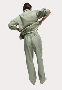 Groene oversized jas met opgerolde mouwen en bijpassende elastische broek. De stof lijkt lichtgewicht met een soepele textuur en subtiele draping.