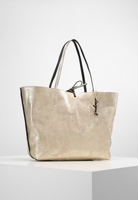 Kultainen metallinen ostoskassi mustilla nahkakahvoilla, jossa on kohokuvioitu kukkakuosi ja koristeellinen riipus. Tilava muotoilu avonaisella päällä.