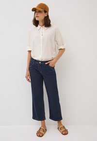 Camicia button-up beige chiaro con maniche arrotolate, jeans a gamba larga blu scuro e cappellino da baseball in suede beige. Sandali intrecciati beige con dettaglio a fibbia.