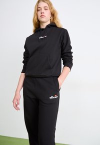 Zwarte hoodie en joggers in zachte stof, met een voorzak, ribbelboorden en een logo op de borst en dij. Eenvoudig, relaxed ontwerp.