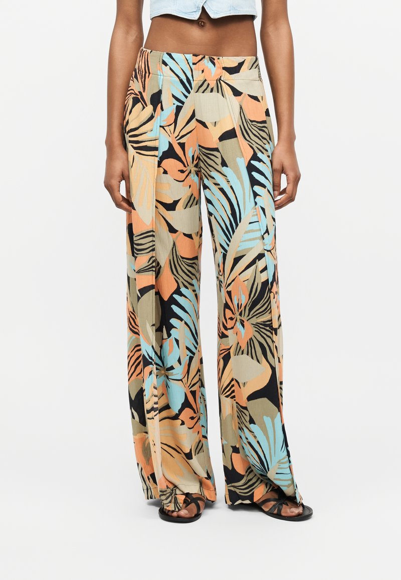 Γυναίκα που φορά ψηλόμεσο, φαρδύ παντελόνι με πολύχρωμο αφηρημένο σχέδιο φύλλων σε ροδακινί, μπλε, μπεζ και μαύρο, συνδυασμένο με μαύρα σανδάλια.