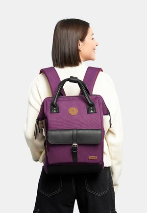 Purple backpack with black leather accents, top handle, adjustable straps, and front flap with a buckle. Features a logo patch.