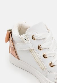 Hvit sneaker med en glatt skinnoverdel, gulløyer, glidelås på siden og rosé gulldetalj på hælen. Snøring og polstret tunge.
