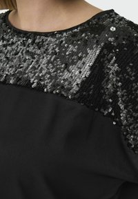 Czarna bluzka z górną częścią pokrytą cekinami i gładką tkaniną poniżej. Okrągły dekolt z subtelnym kontrastem tekstur. Brak widocznego logo.