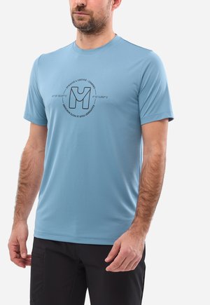 Mann trägt hellblaues Kurzarmhemd mit rundem Logo und schwarze Hose, steht vor einem schlichten weißen Hintergrund.