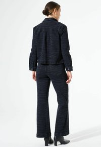 Veste en denim et pantalons évasés assortis en bleu foncé avec un motif ondulé subtil, présentant un col et des détails de boutons, portés avec des bottes à talons.