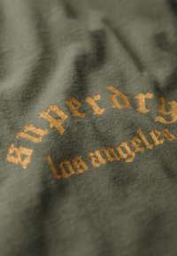 Tejido verde oliva con una textura suave que presenta letras doradas que dicen "Superdry los ángeles". Sutiles arrugas añaden profundidad al material.