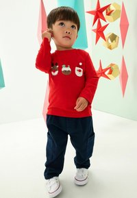 Chemise rouge à manches longues avec trois personnages de vacances brodés, assortie à un pantalon cargo bleu marine et des baskets blanches.