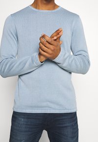 Hellblaues Langarmshirt mit gerippter Textur. Es hat einen runden Ausschnitt und sichtbare Nähte entlang der Nähte. Wird mit dunklen Jeans getragen.