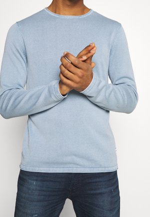 Hellblaues Langarmshirt mit gerippter Textur. Es hat einen runden Ausschnitt und sichtbare Nähte entlang der Nähte. Wird mit dunklen Jeans getragen.