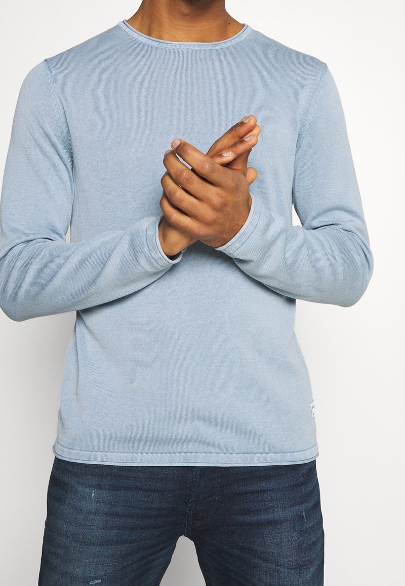 Hellblaues Langarmshirt mit gerippter Textur. Es hat einen runden Ausschnitt und sichtbare Nähte entlang der Nähte. Wird mit dunklen Jeans getragen.