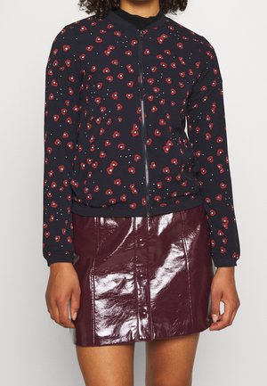 Femme portant une veste noire zippée avec des motifs floraux rouges et une jupe en cuir bordeaux brillante à boutons.