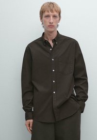 Giovane con capelli biondi corti, indossa una camicia button-up marrone scuro e pantaloni abbinati su uno sfondo chiaro e semplice.
