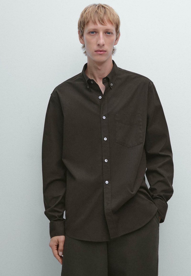 Giovane con capelli biondi corti, indossa una camicia button-up marrone scuro e pantaloni abbinati su uno sfondo chiaro e semplice.
