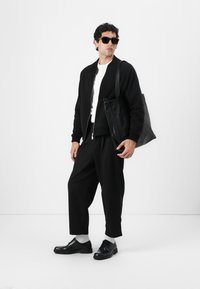 Chaqueta bomber negra sobre una camiseta blanca, pantalones negros holgados, zapatos negros, calcetines grises y bolso de mano de cuero negro. Se llevan gafas de sol oscuras.