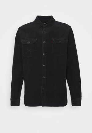 Chemise noire en velours côtelé à manches longues avec fermeture à boutons, deux poches poitrine à rabat boutonné et col pointu sur fond blanc.