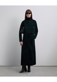 Veste courte en velours côtelé vert foncé avec deux poches avant, associée à une jupe longue assortie. Le modèle porte des lunettes de soleil noires et des bottines.